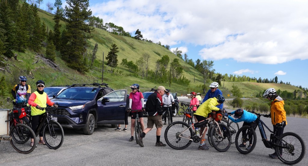 A group of people with bicycles and cars parked on a gravel road

Description automatically generated