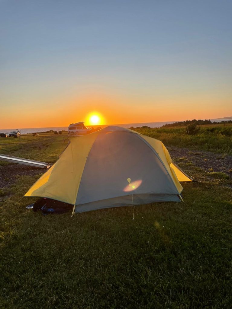 A tent on grass with the sun setting in the background

Description automatically generated
